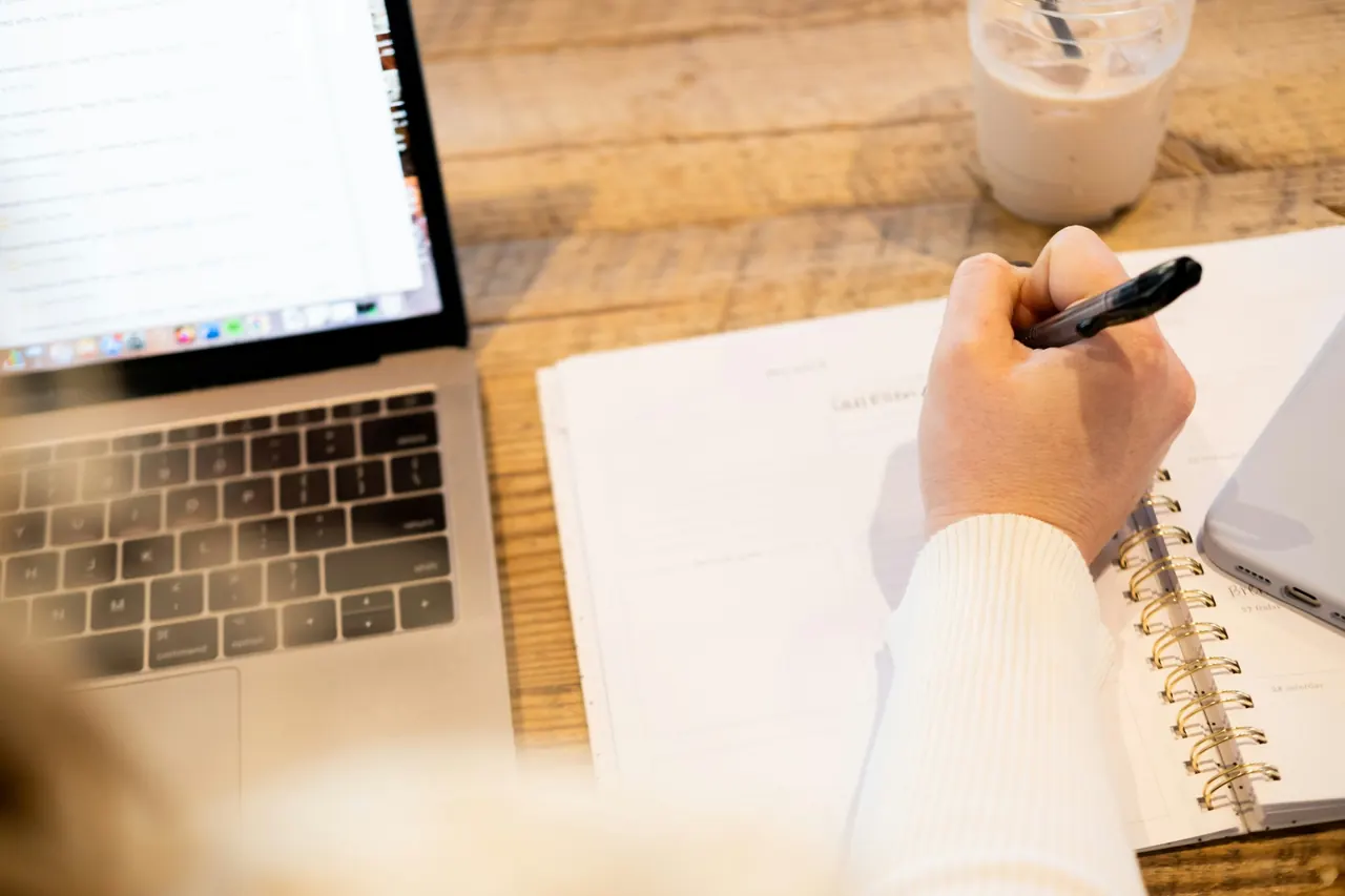 Over-the-shoulder view of someone writing in a planner beside an open laptop